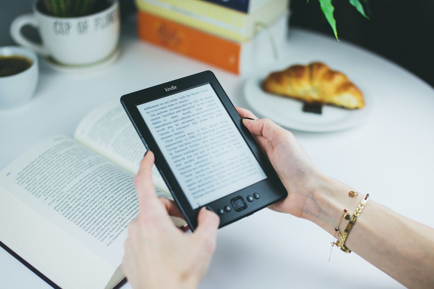 Two hands holding a Kindle with a physical book in the background