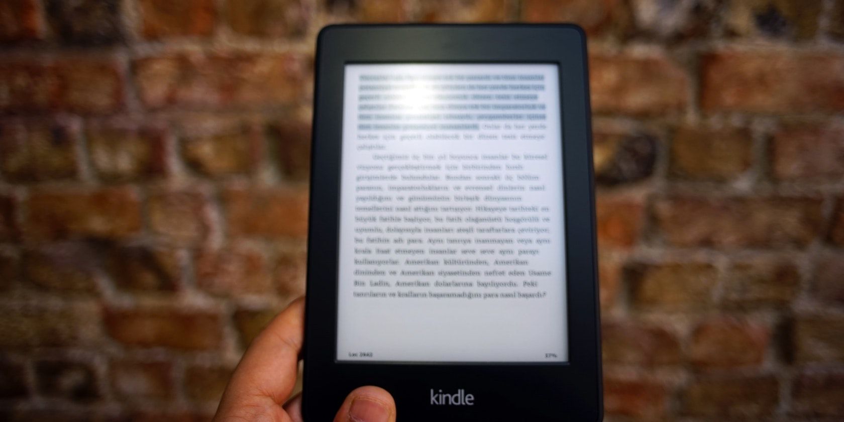 A hand holding a Kindle in the foreground with a brick wall in the background
