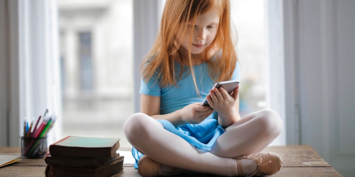 Crossed legged ginger girl in blue dress, holding phone next to stack of books and pens