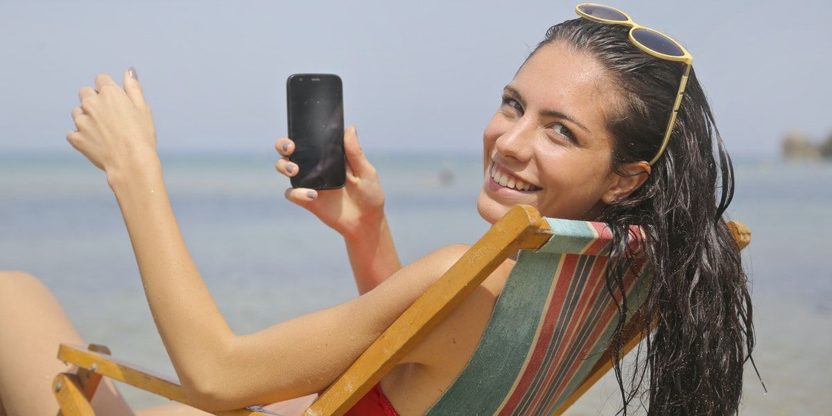 Woman hald turned in chair at beach smiling at camera and holding phone