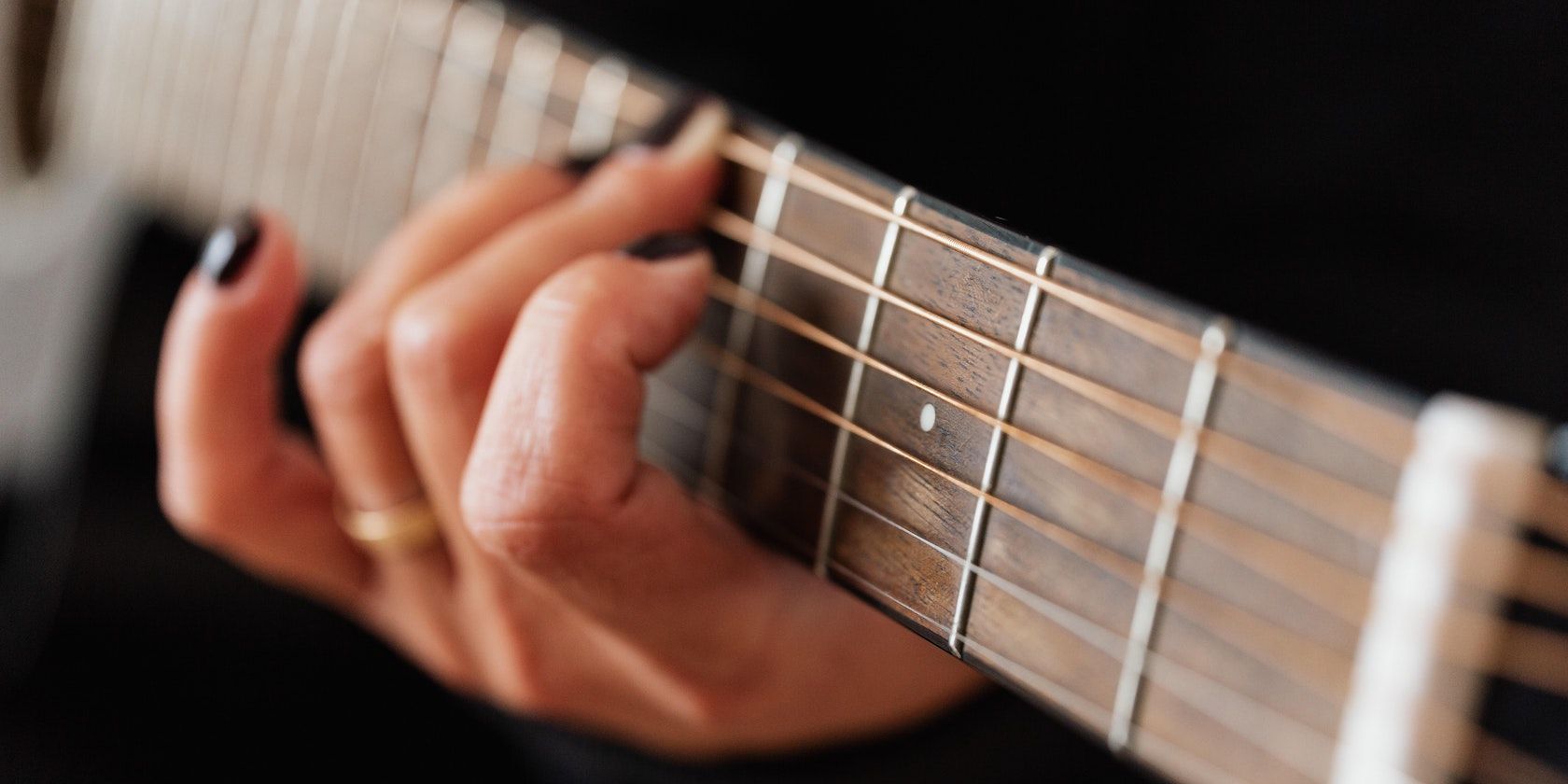 A focused shot of a hand with painted nails and wearing a ring, positioning on the neck of a guitar.