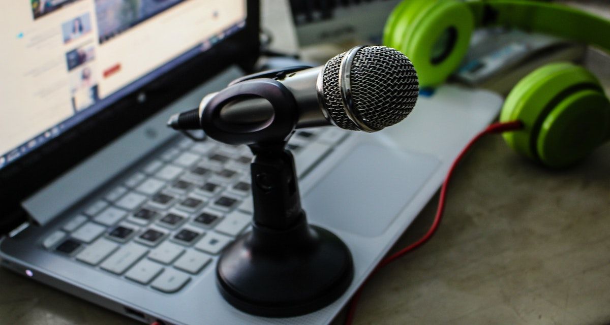 A microphone on a laptop with a pair of green headphones in the background.