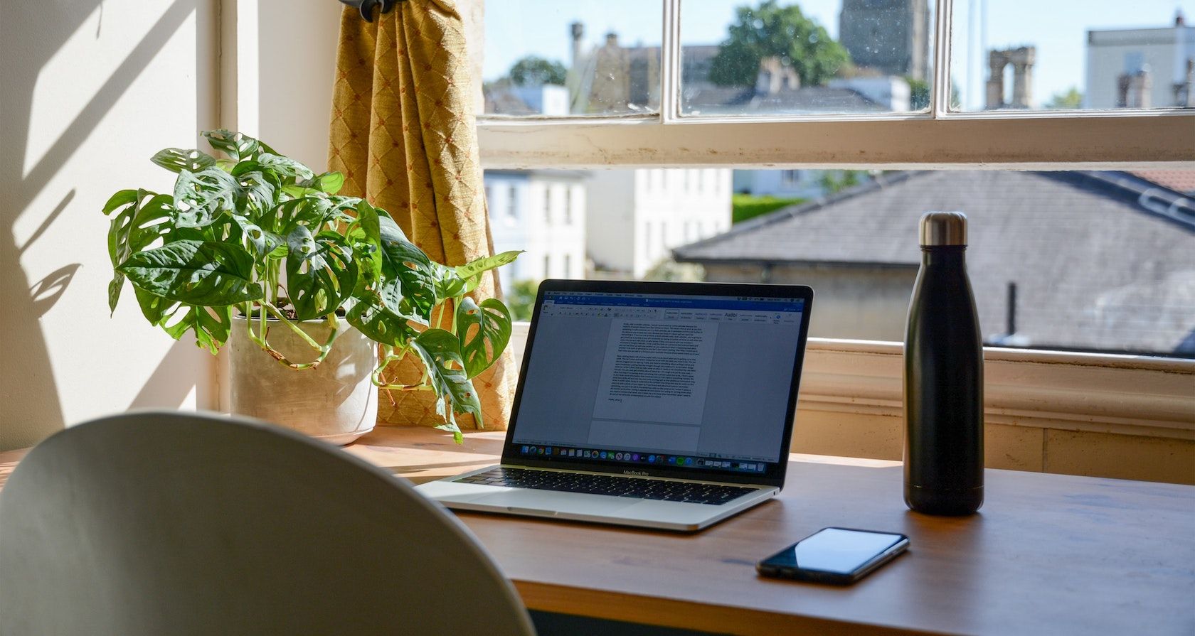 A student's Macbook, iPhone, water bottle, and plant on their desk by a window
