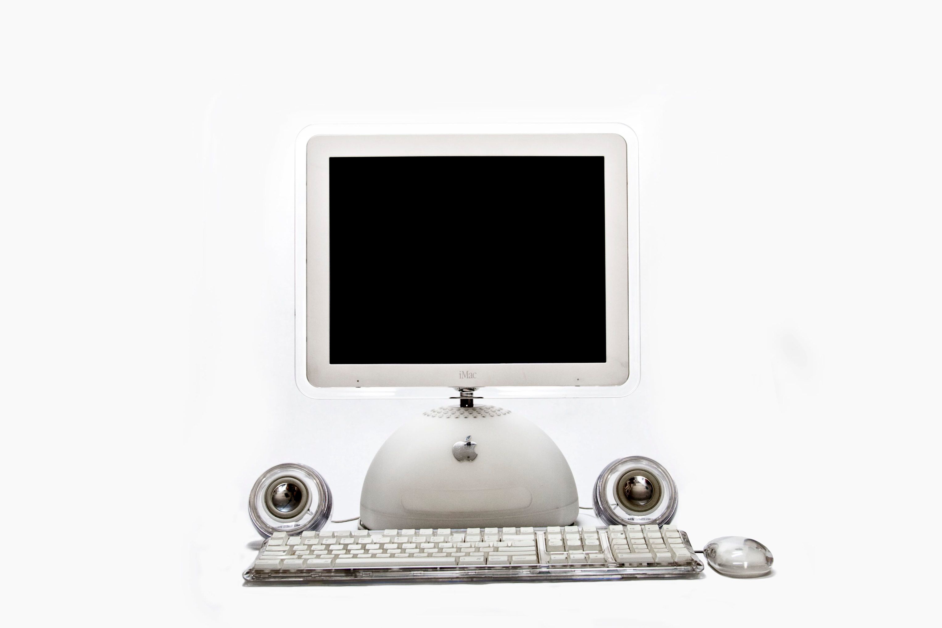 An iMac G4 sits against a white background with speakers, a keyboard, and a mouse surrounding it