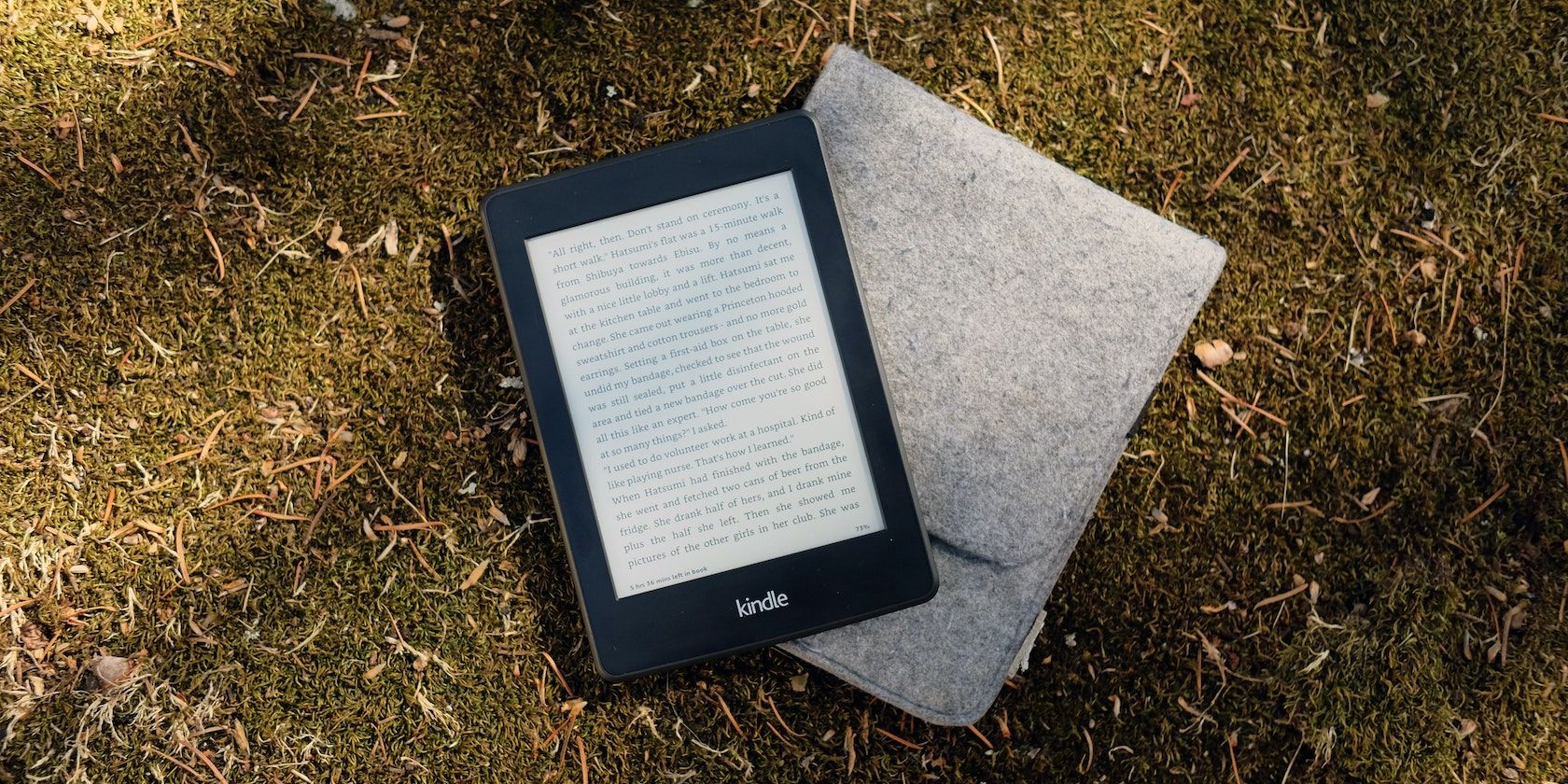 A Kindle on top of a gray flip cover case on grass.