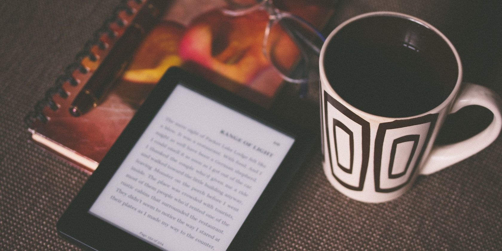 A Kindle on top of a notebook and next to a black and white mug glasses and pen