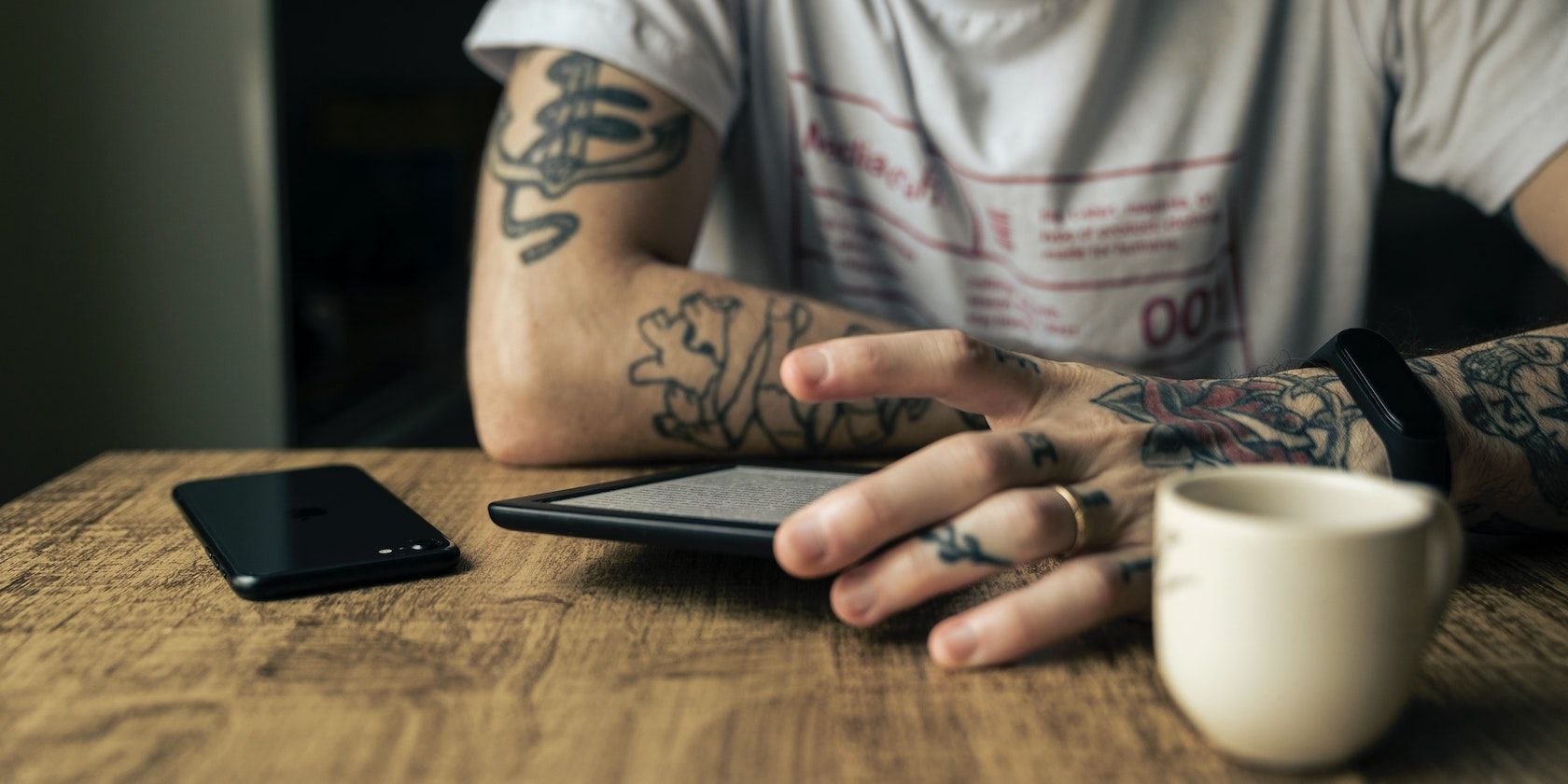 A person with tattoos reading a Kindle on a wooden table with a black phone and white mug beside it