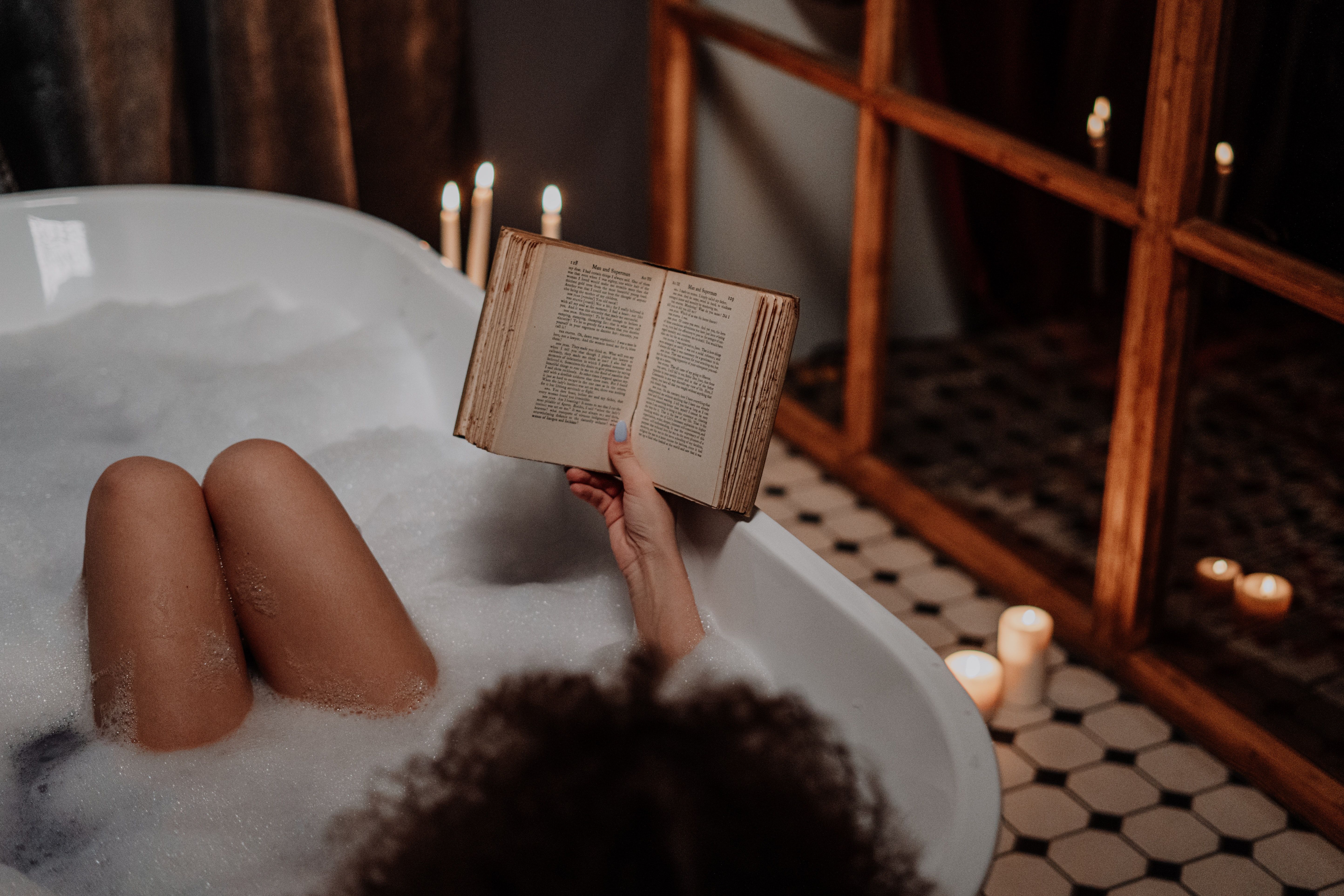 Woman sitting in a bathtub reading a book among scented candles