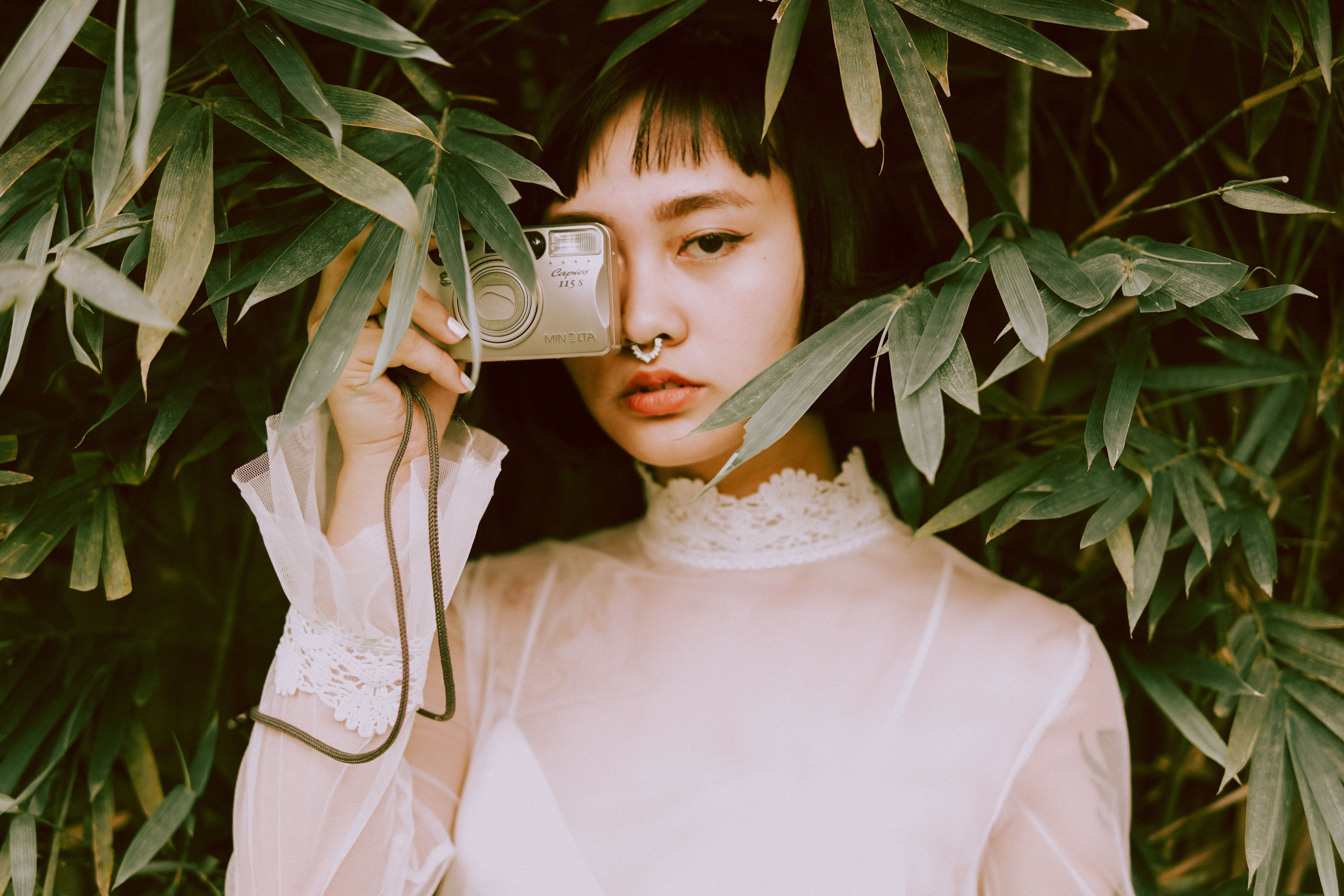 A young woman standing behind leaves and covering of her piece of her eye with a camera