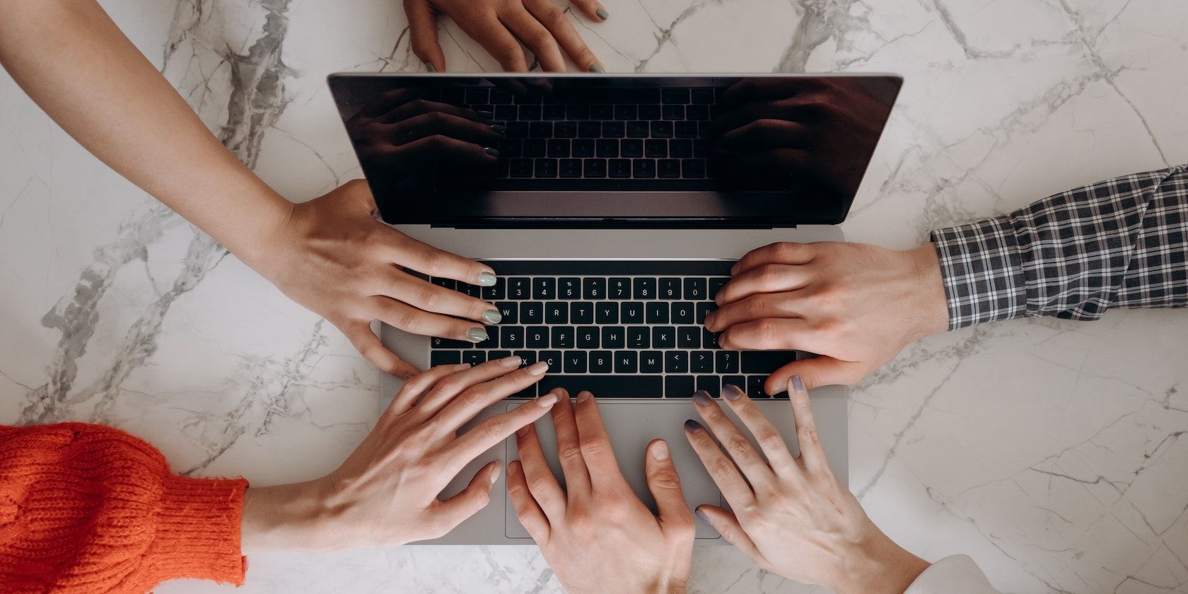 Multiple hands from multiple people all touch different parts of a MacBook keyboard.