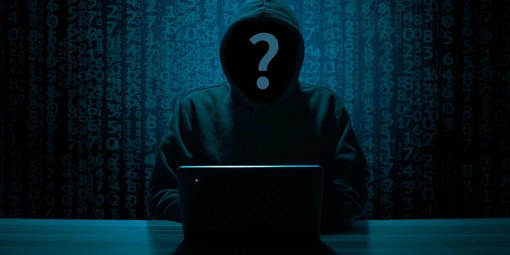 Anonymous hacker's face in a black hoody with a PC on the table