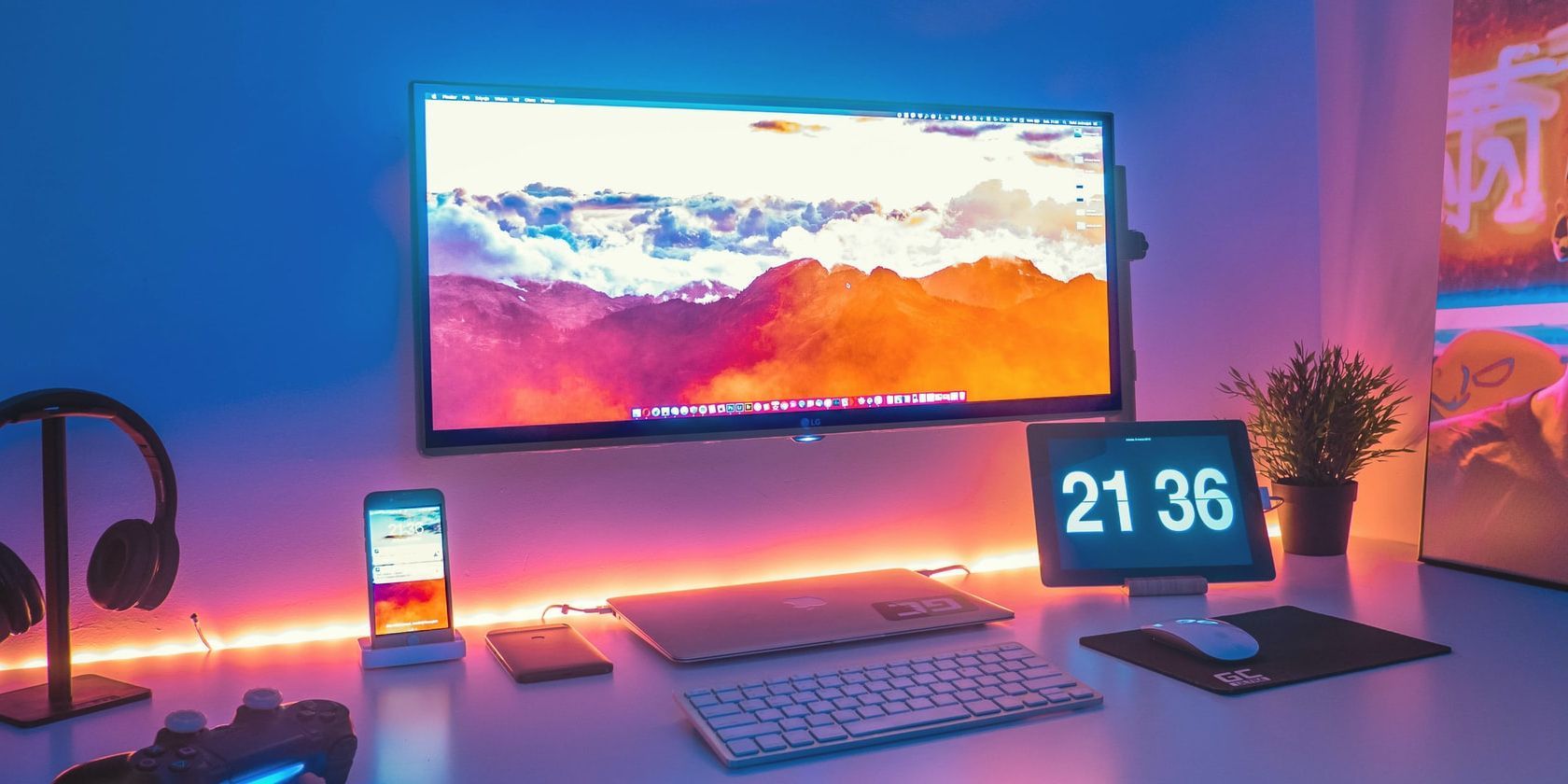 Wall mounted monitor with clouds and mountains desktop background above a desk holding multiple devices. Mood lighting resembles a sunset.
