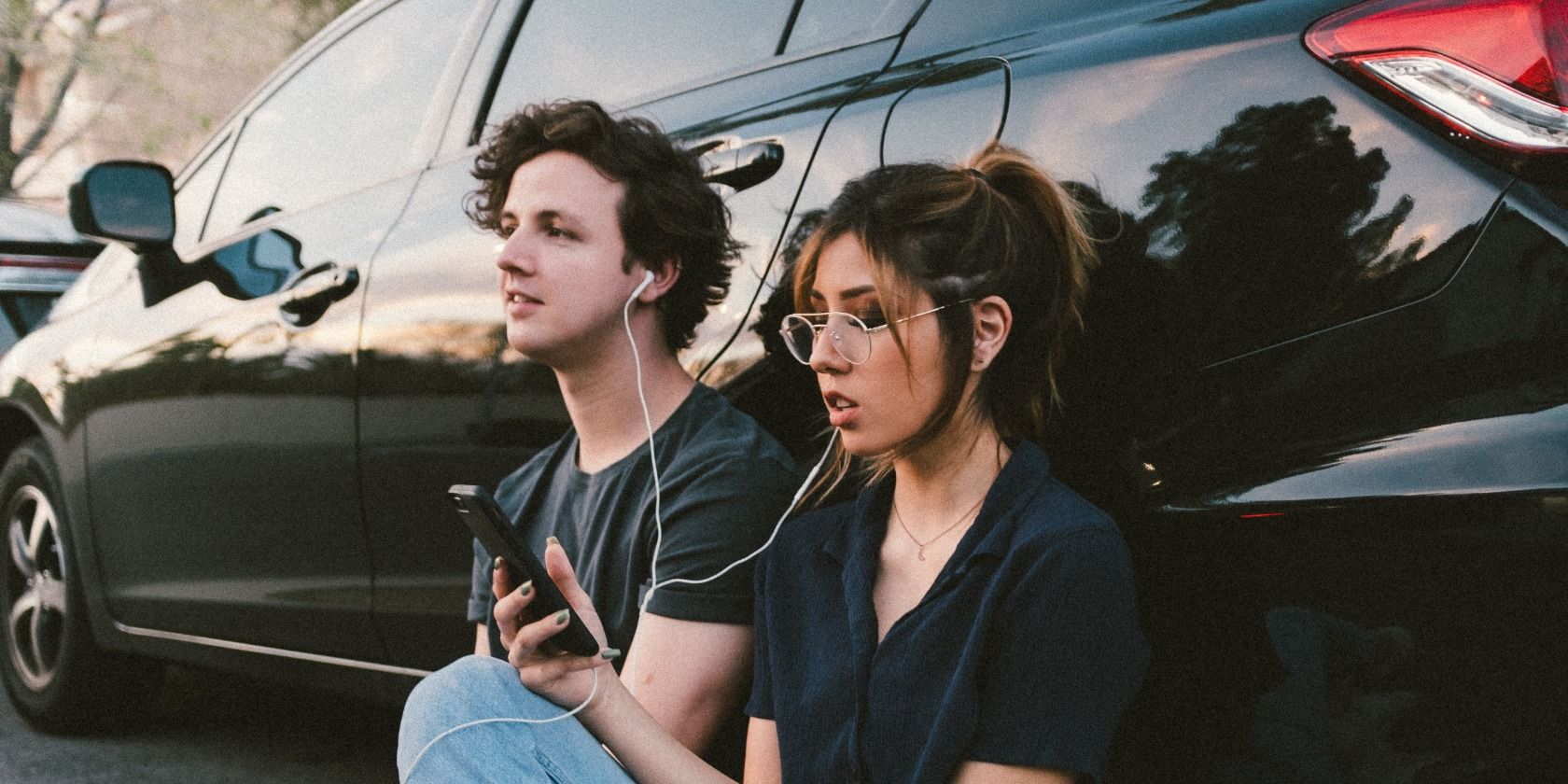 young man and woman using earphones, listening to music against a car
