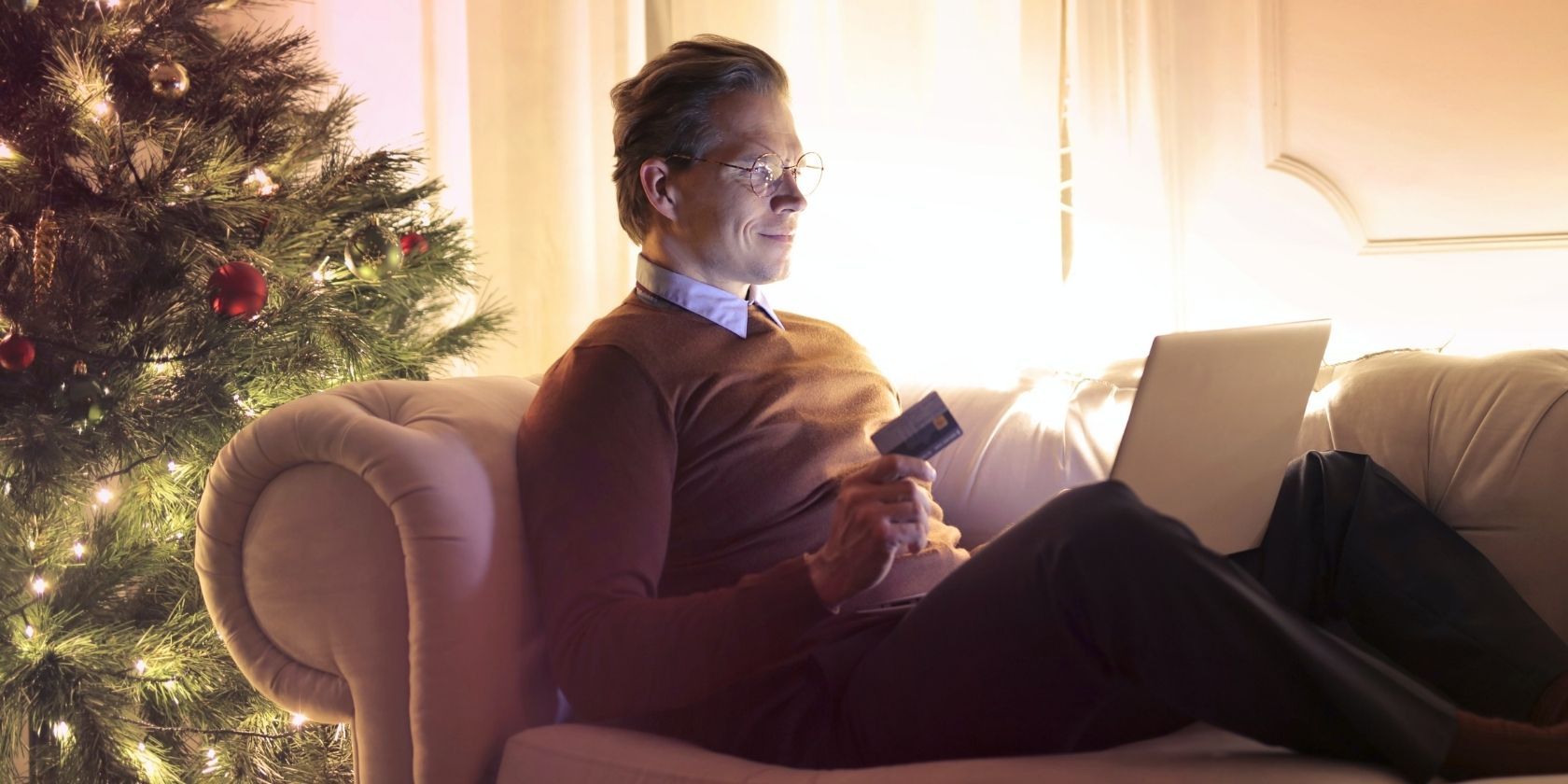 Man near Christmas tree with laptop laying on couch with a credit card in his hand