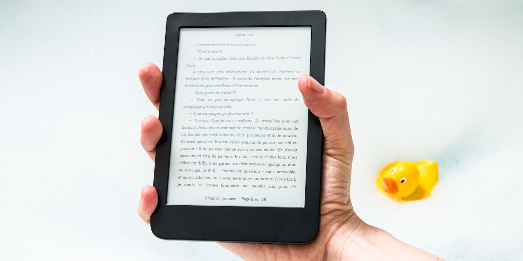 A close up shot of a hand holding an e-reader on a white-blue background with a yellow rubber duck in the background