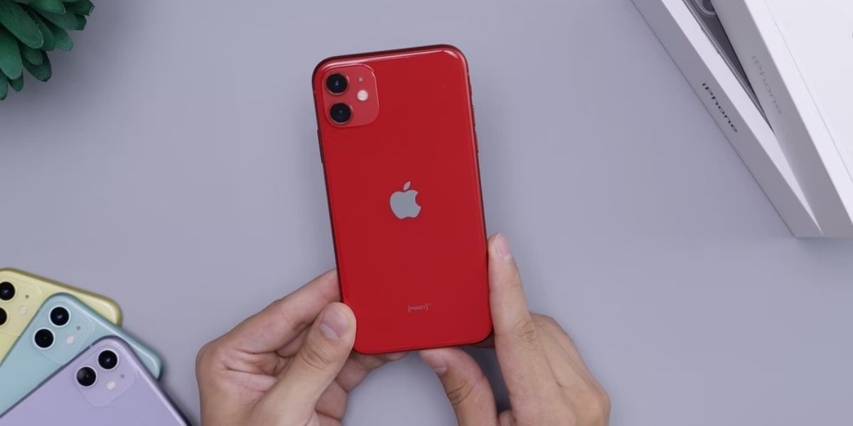 hands holding red iphone 11 on white desk with yellow, green, and purple iphone 11 in the corner
