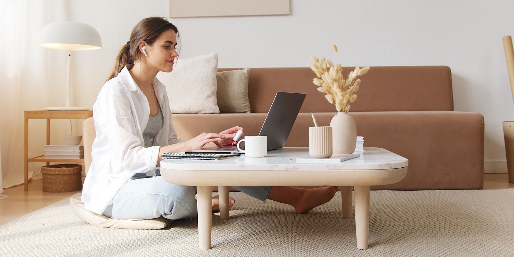 A woman sitting on the floor typing on her laptop at the coffee table. The room is clean and tranquil with lots of beige colors