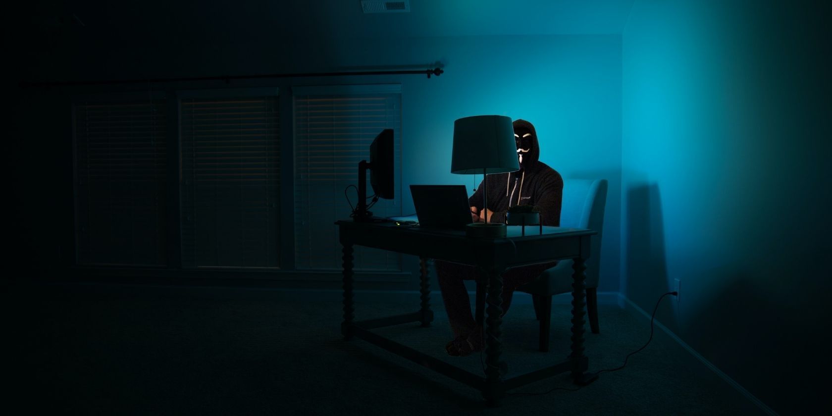 Masked man in all black sitting at computer in a dark, blue screen-lit room
