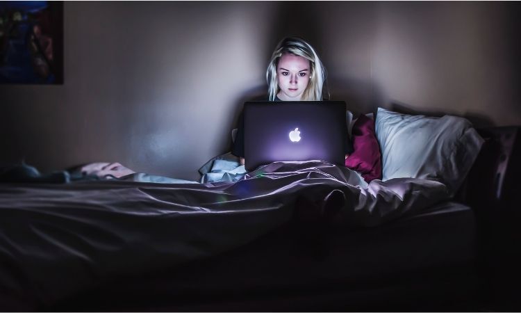 Woman in bedroom on bed lit by her laptop screen