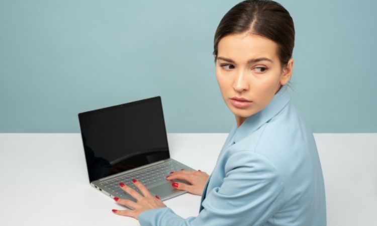 brunette woman in blue blouse with blue background working on laptop and looking over shoulder