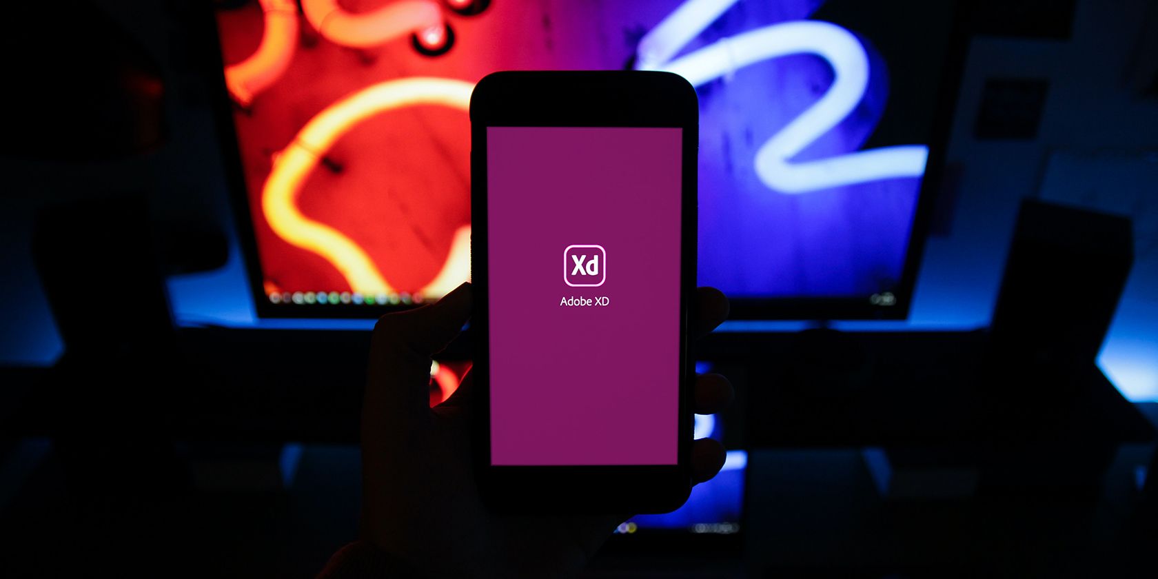 A dark photo of a mobile phone showing the Adobe XD home screen, background consists of lit neon colors on a desktop screen.