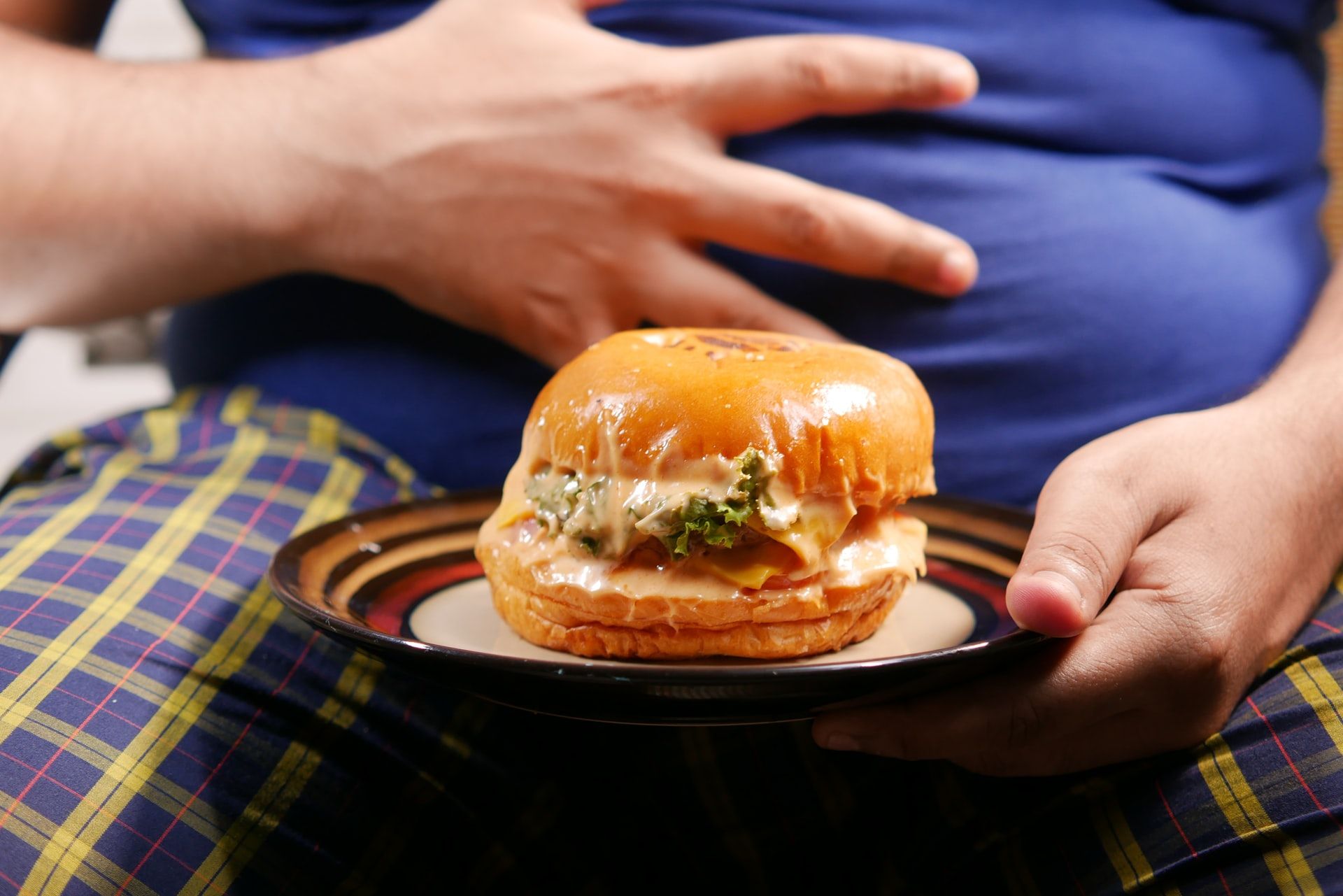 Fat Man Placing One Hand on Belly While Holding Burger In A Plate
