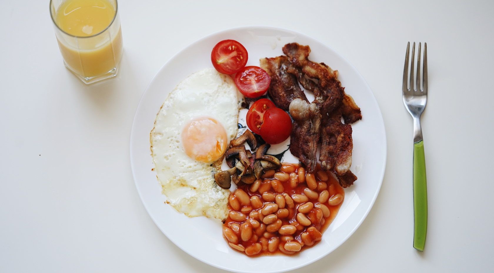 plate of bacon eggs and beans on table next to fork and juice