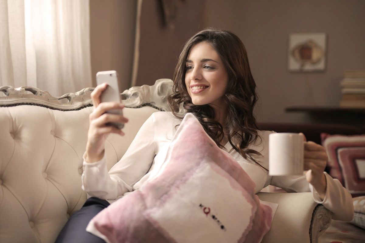 smiling woman in white long sleeved shirt staring at phone on sofa