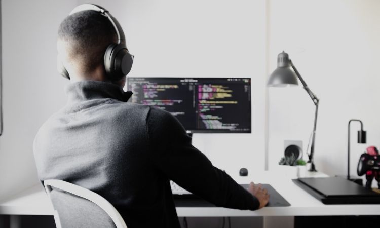 man in long black sweater wearing white headphones and typing coding on computer