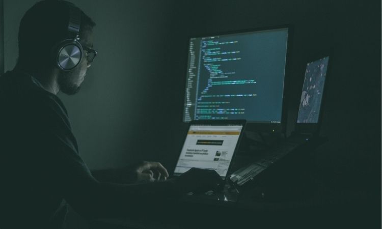 man with headphones on sitting in dark room lit by computer screen with coding