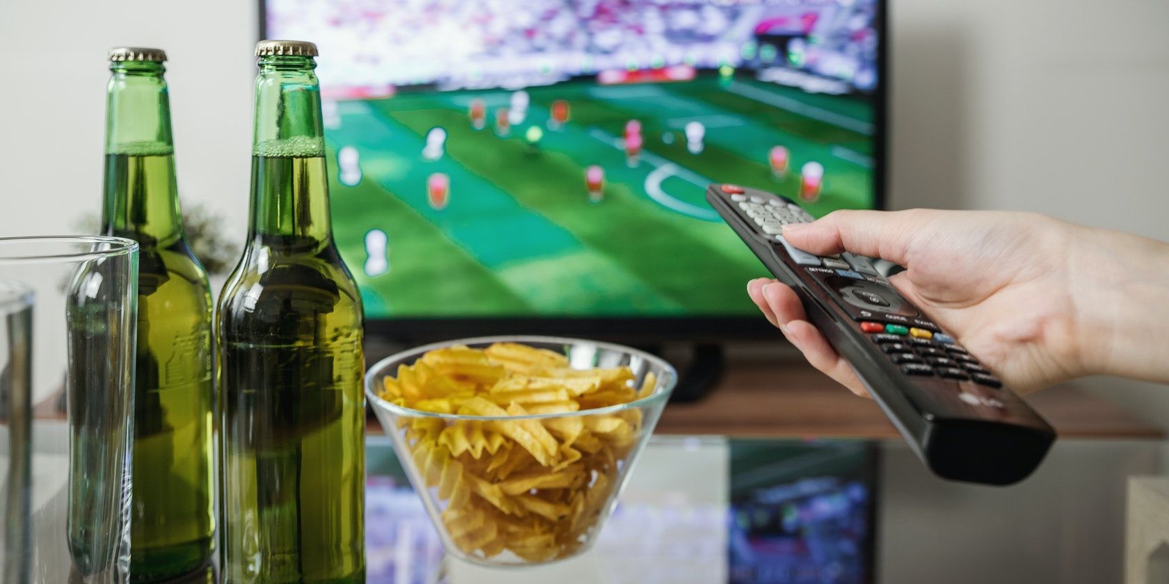 Des collations, des boissons et une main tenant une télécommande devant un écran de télévision diffusant un match de football