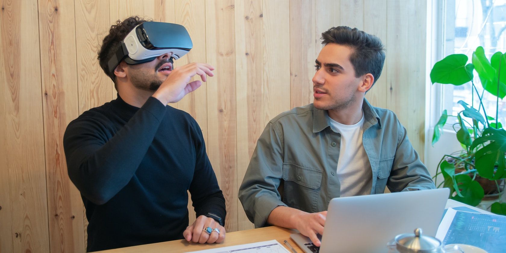 Image of a man using a virtual reality device while sitting next to another man using a laptop