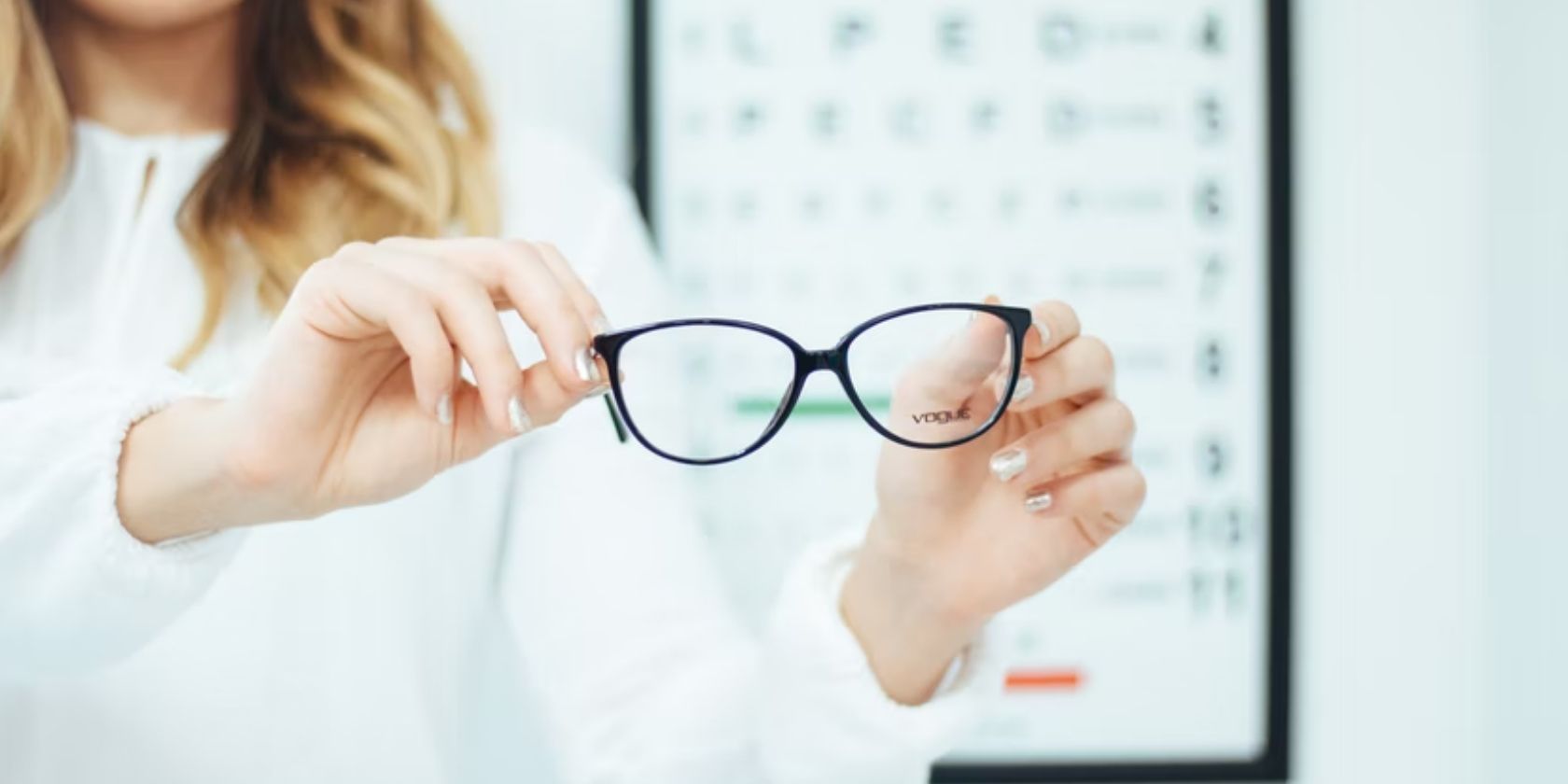 woman holding a pair of black framed glasses with eye test board in background