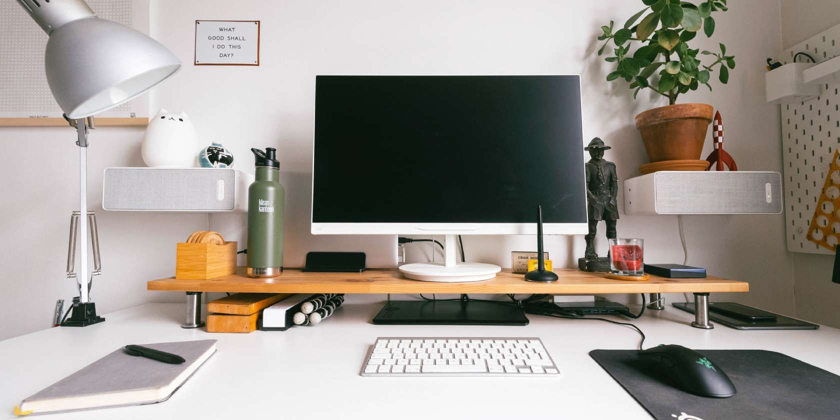  A person is working at a desk with a computer, keyboard, mouse, and notebook. The desk is organized with a lamp, plant, speaker, and other items on it.