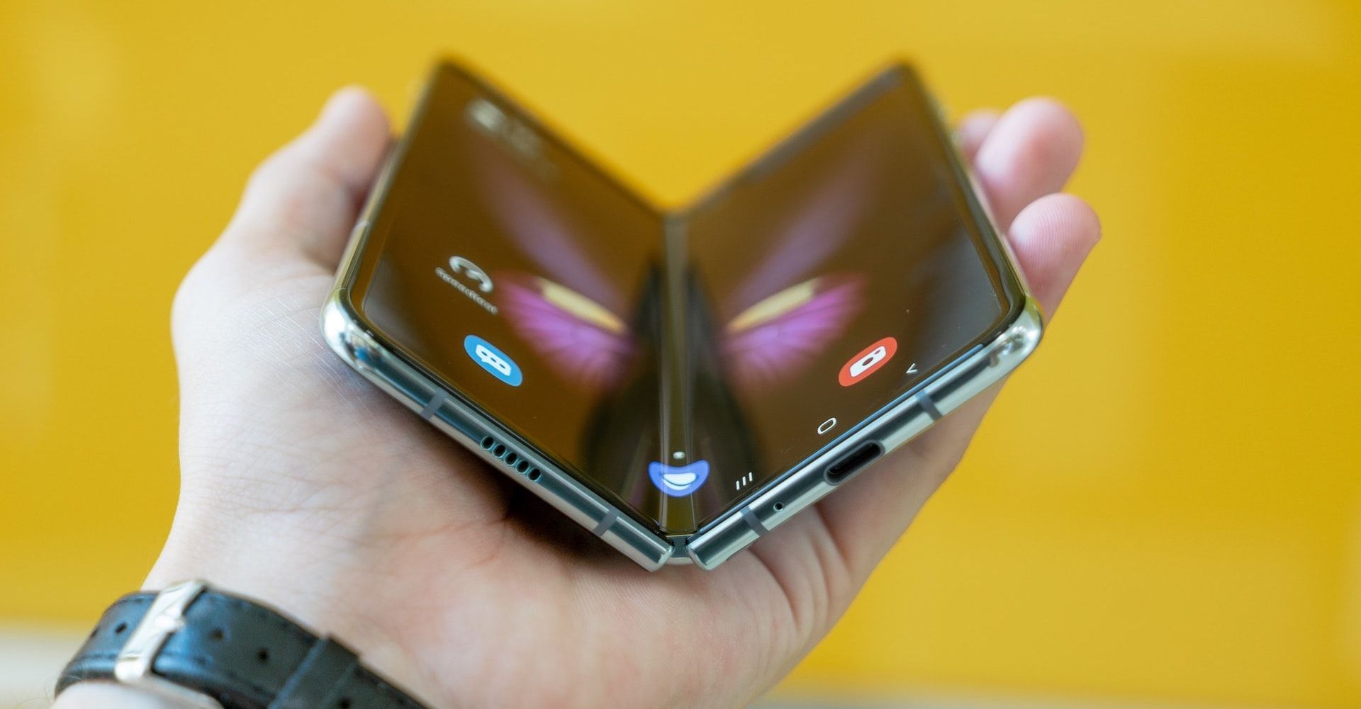 A single hand holding a Galaxy Fold phone half open against a bright yellow background