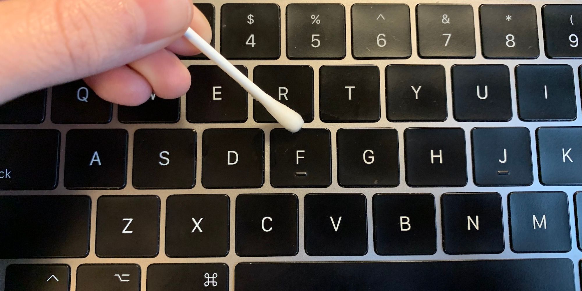 A hand holding a cotton swab wiping inbetween keys on a macbook keyboard