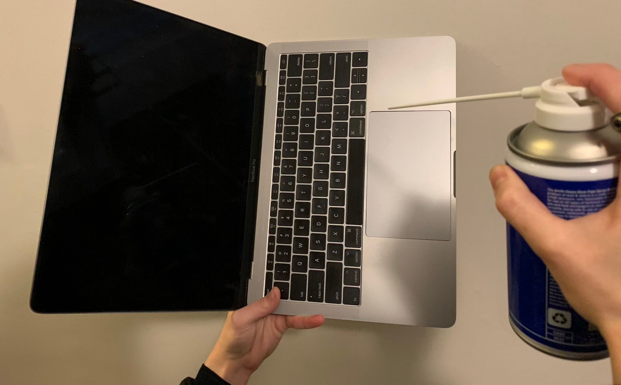 A hand holding an open macbook in the air, rotated 90 degrees to the left, with a hand pointing a compressed air can at the keyboard