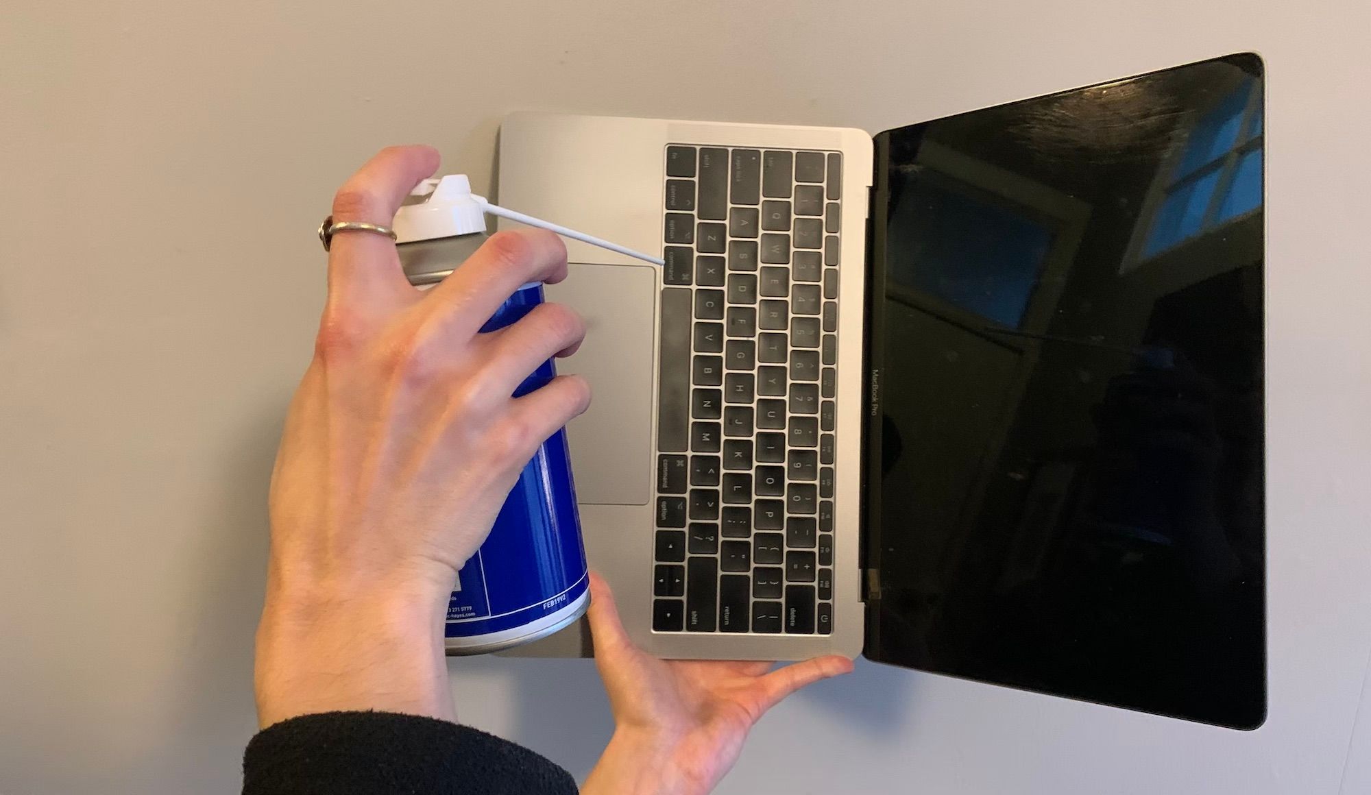 A hand holding an open macbook in the air, rotated 90 degrees to the right, with a hand pointing a compressed air can at the keyboard