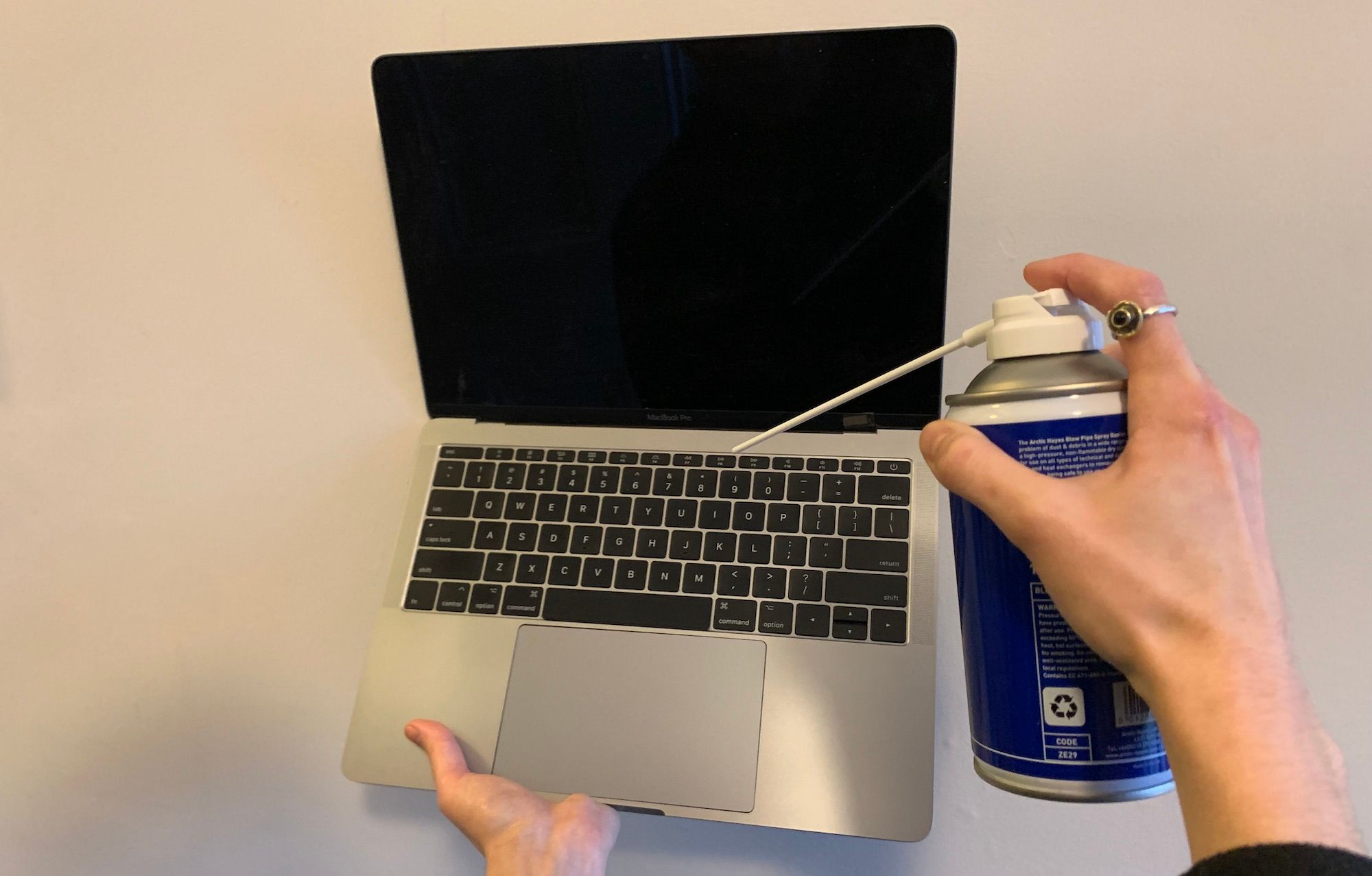 A hand holding a macbook in the air with a hand pointing a compressed air can at the keyboard