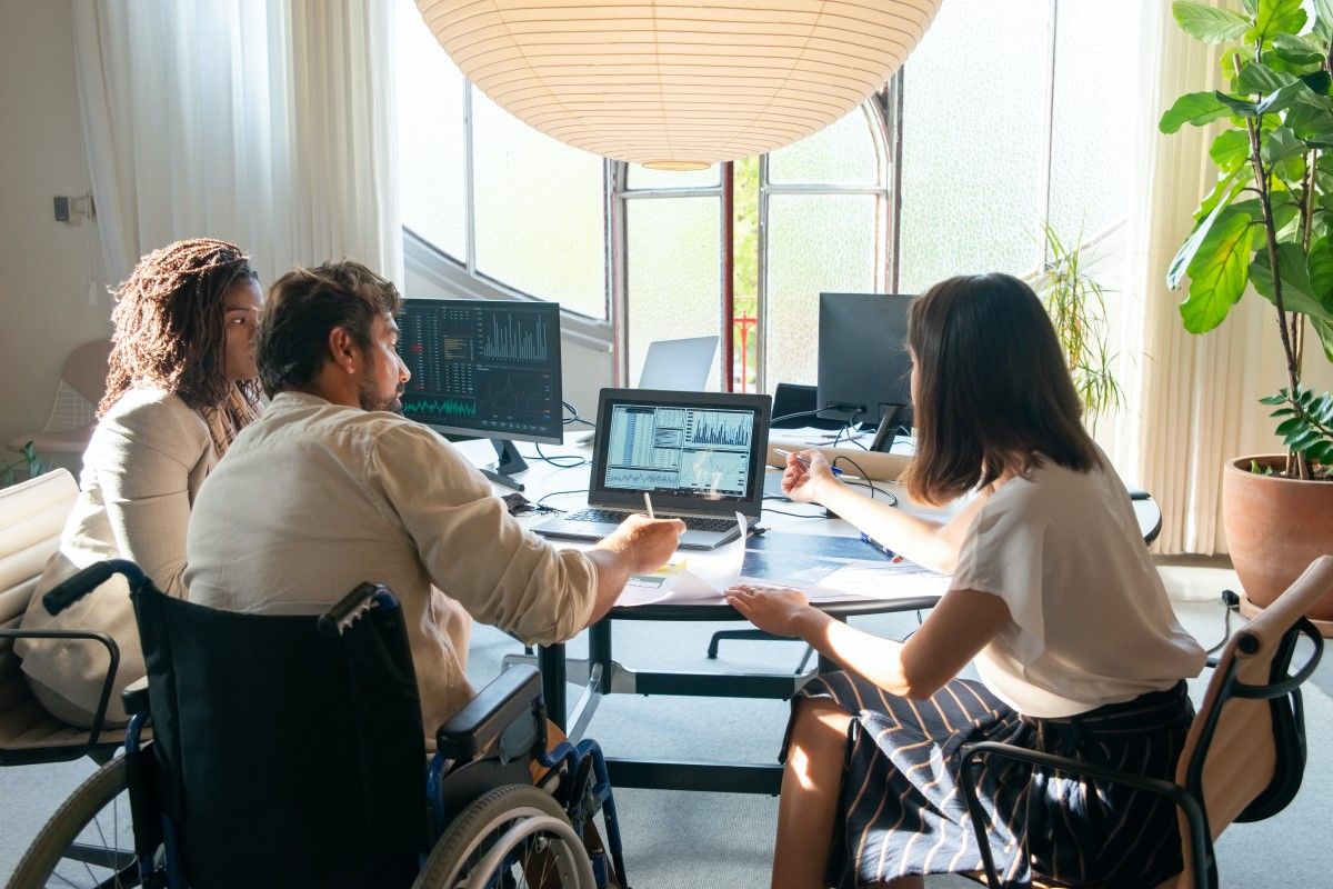 Three people work around a table in an office. The man in the center is in a wheelchair and there are women on both sides of him. They are looking at a computer with financial forecasts on it. 