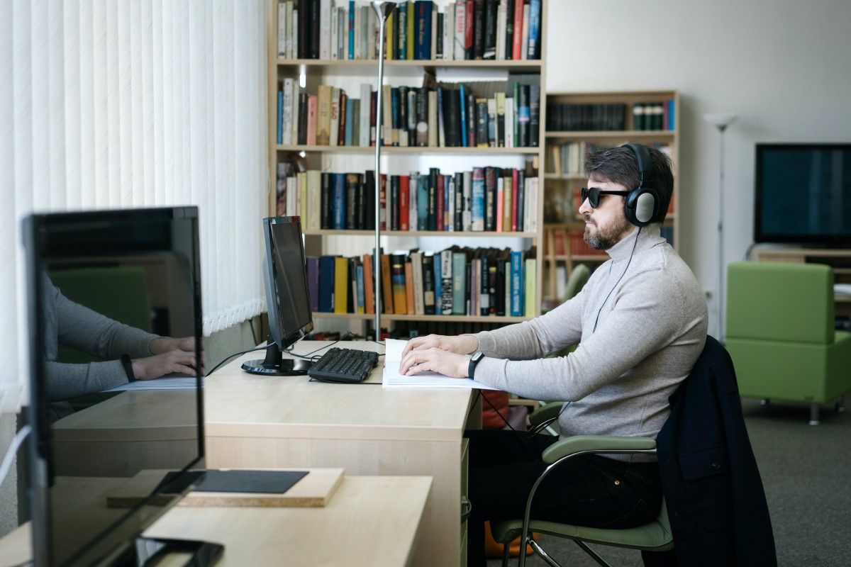 A blind man sits at a computer in a library with dark glasses on