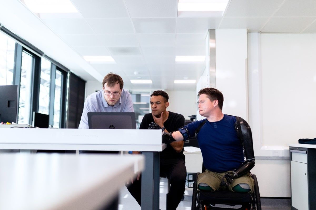 Three men are in an office crowding around a computer. One is in a wheelchair and has lost both legs and an arm. He has a bionic limb