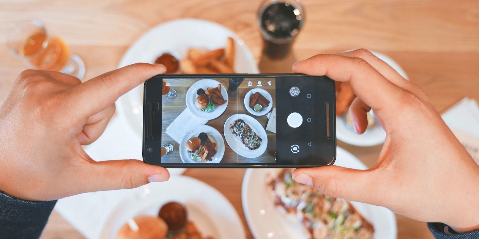An overhead shot of two hands holding a smarphone taking a photo of several plates of food