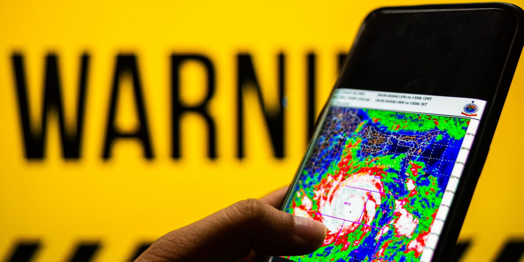 Photograph of a mobile phone with cyclone moving towards the coast shown. The background is yellow with warning sign