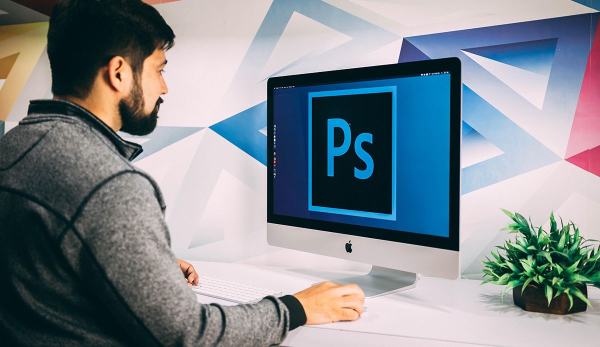 A man sat at a Mac opening Adobe Photoshop on screen.