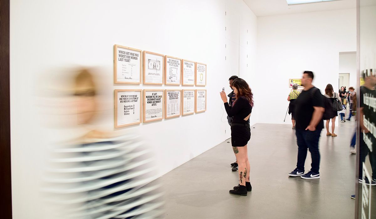 10 framed pieces of typographic design on a wall while people dressed in black look and take photos of them.