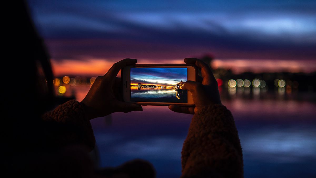 Image of girl holding smartphone taking a photo of the sunset.