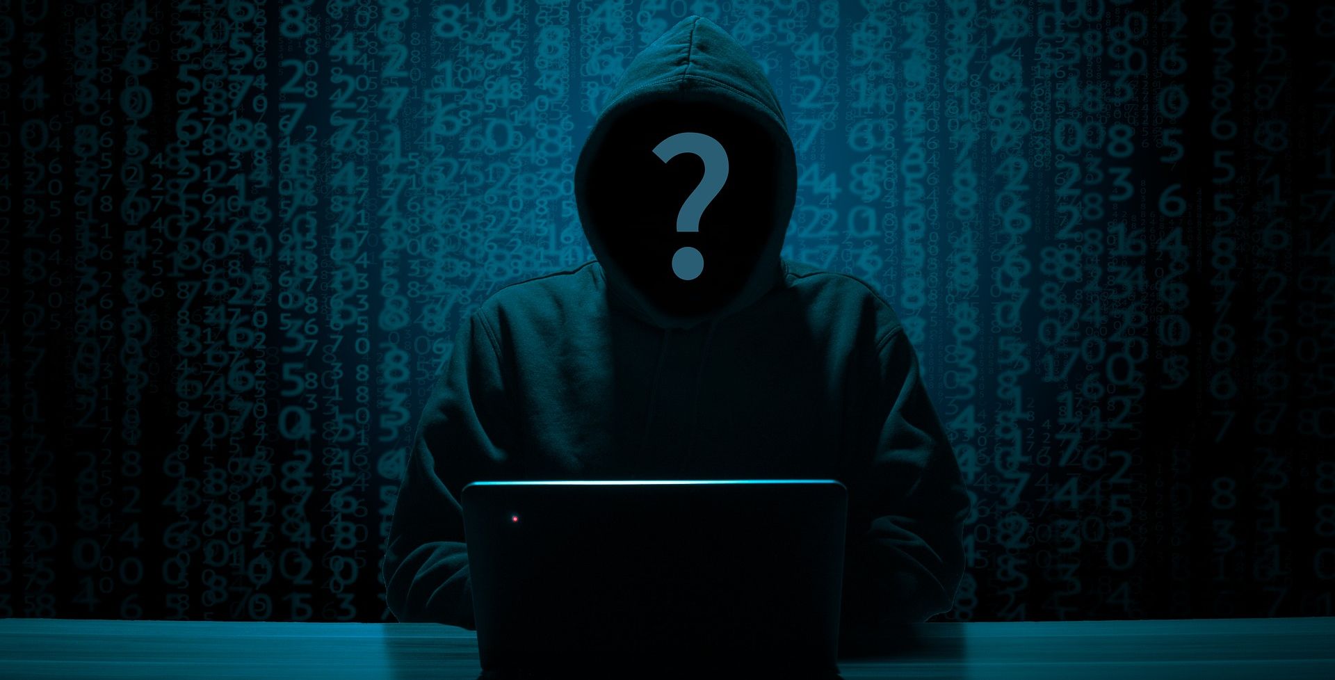 anonymous hooded individual using laptop in front of code background 