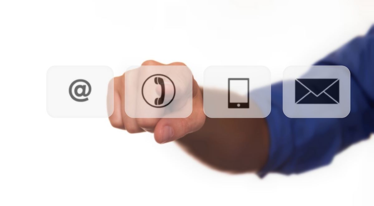 Image of @ Sign, Telephone, Mobile, and Email Icons. Persons Arm and Hand Shown Pointing in the Background. 