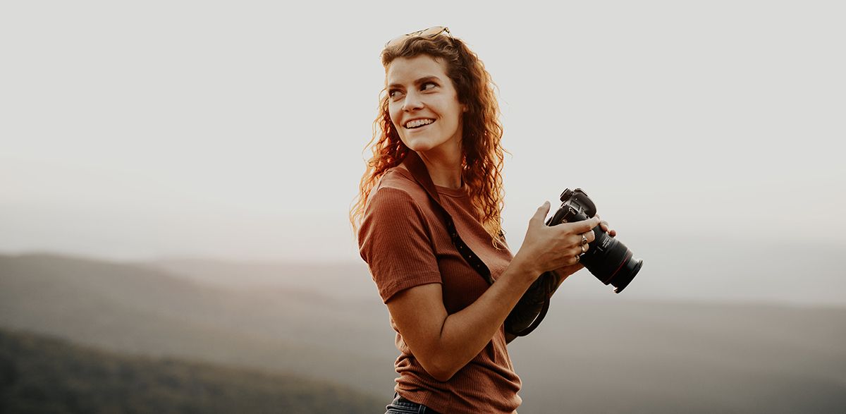 A curly-haired ginger woman holding a DSLR and looking over her shoulder behind her.